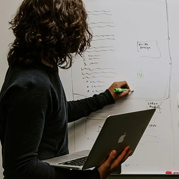 A woman holding a laptop writes notes on a whiteboard
